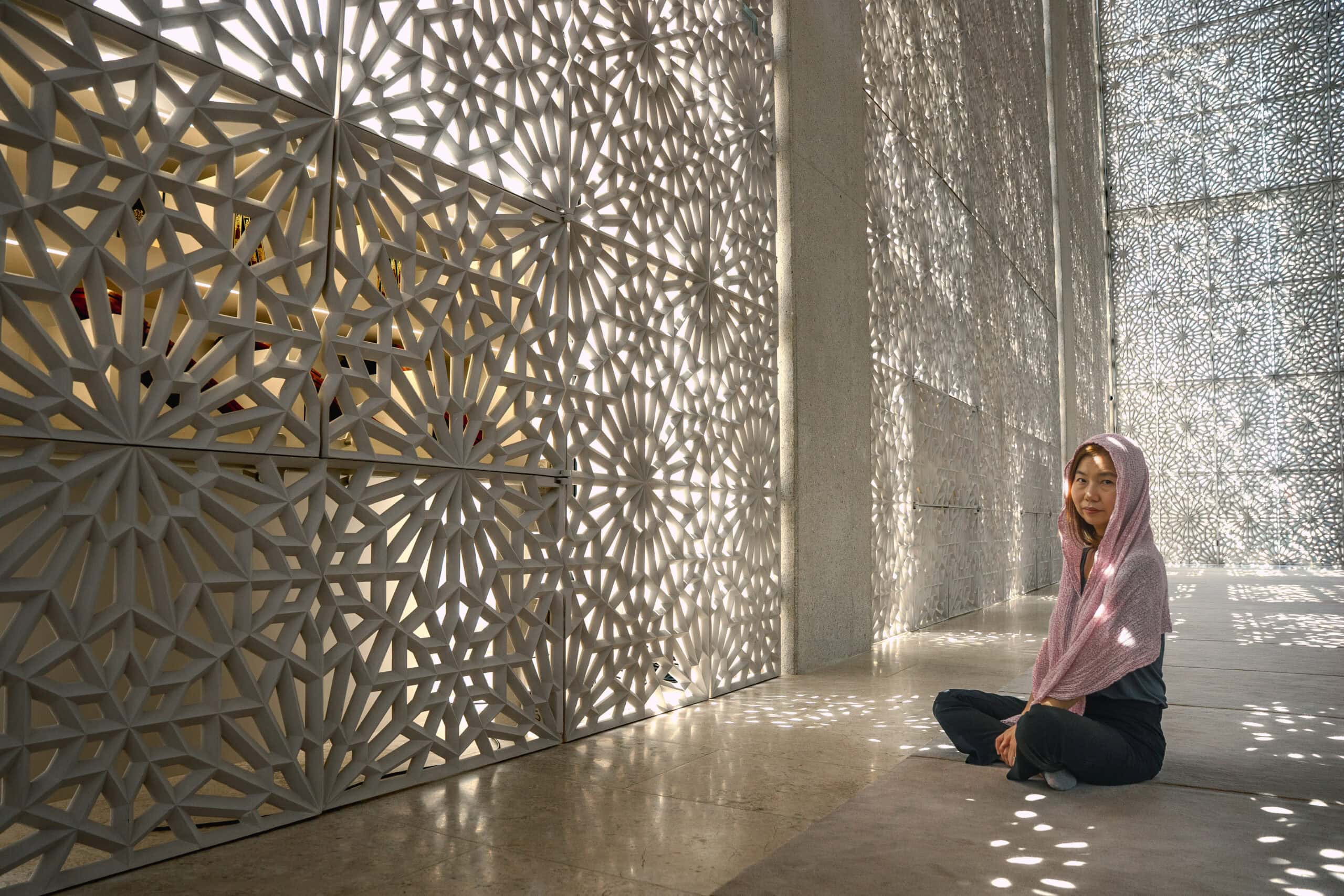 Jin sits on the carpet of the Imam Al Tayeb Mosque, head covered with a scarf. Light is streaming in beautifully though the intricate geometric lattice of the mashrabiya screens