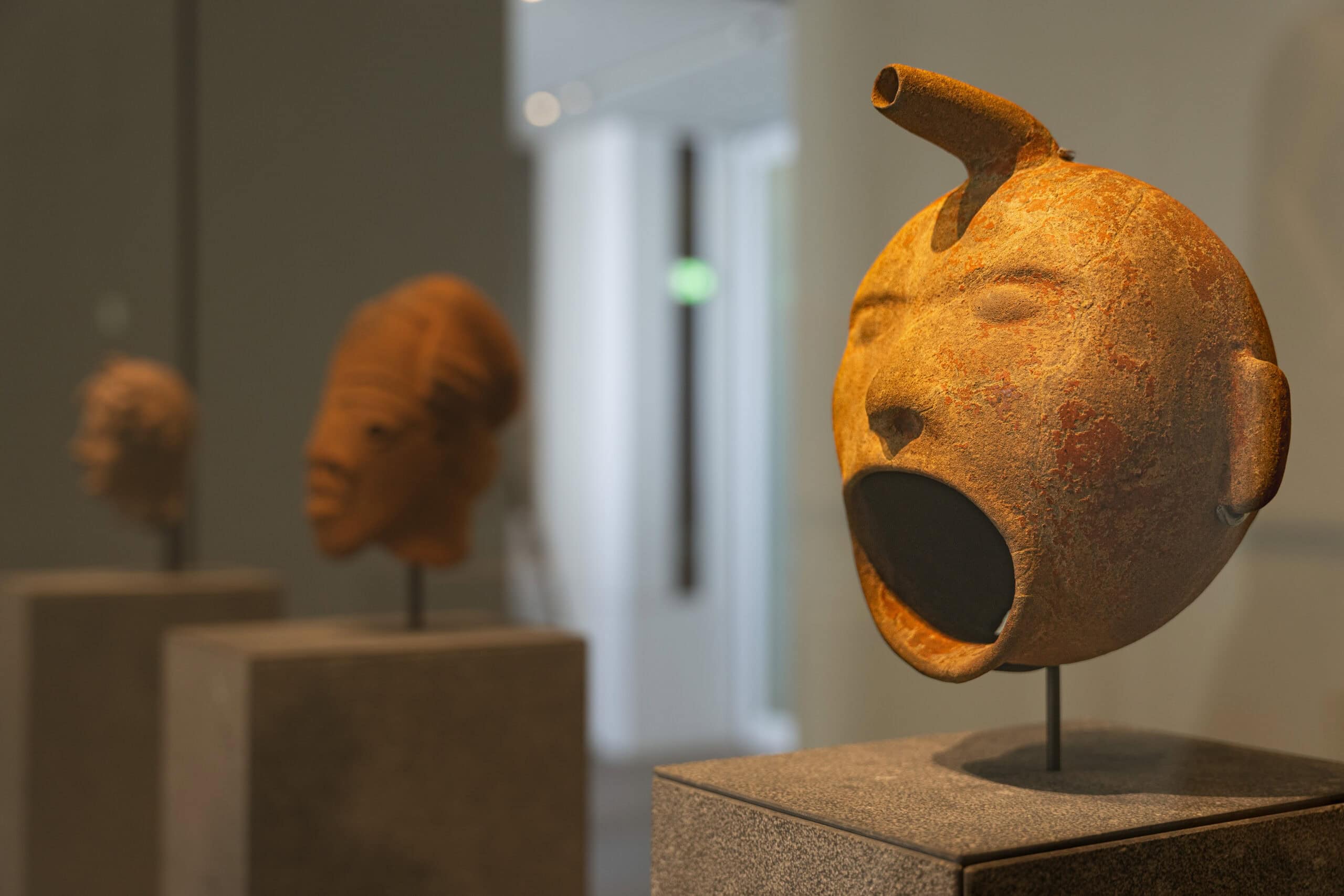 A terracotta mayan vessel that looks like a face with a wide open mouth and a spout for hair