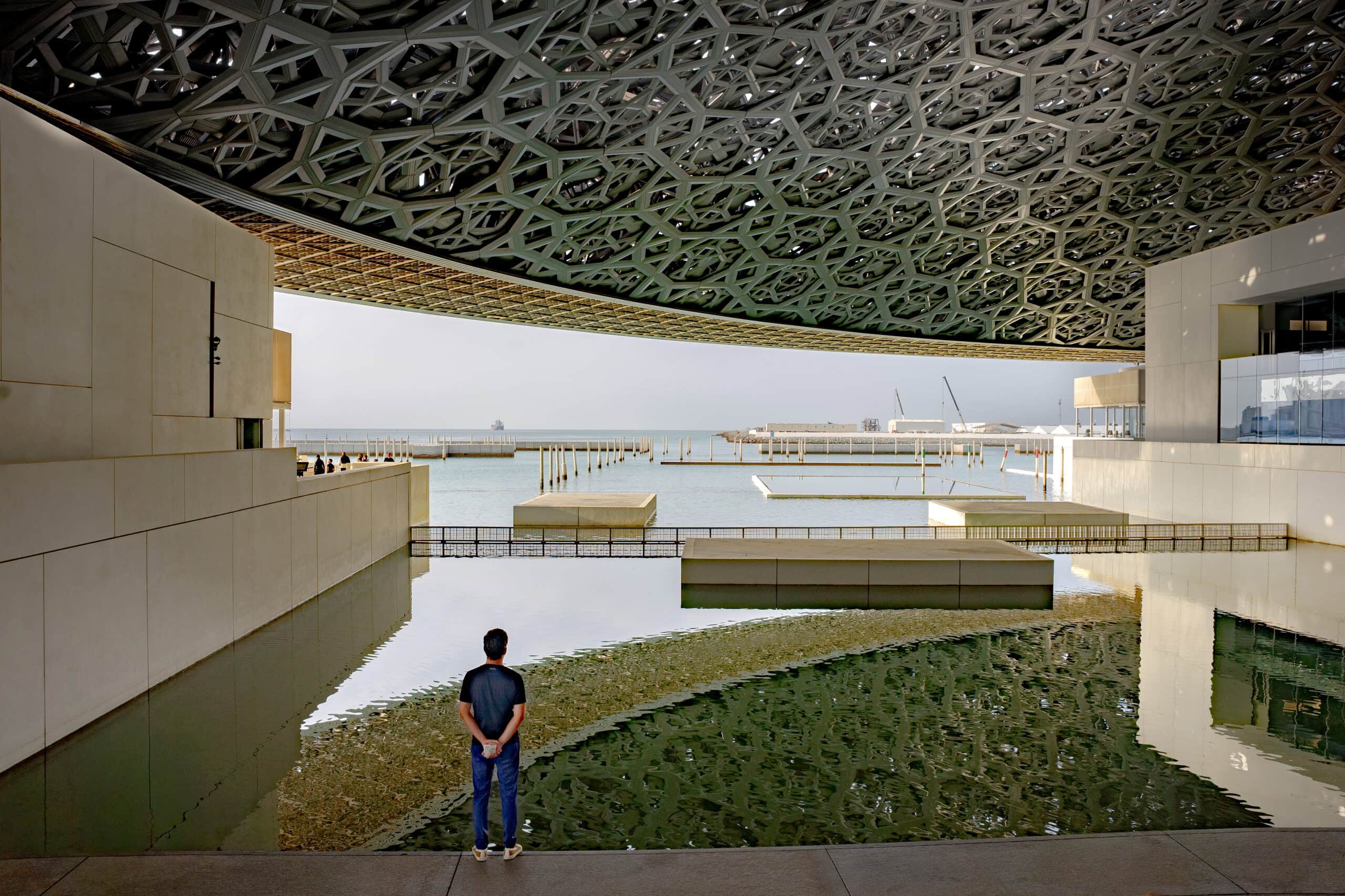 A man stands looking out over the water with the dome of the Louvre Abu Dhabi reflcted in the water