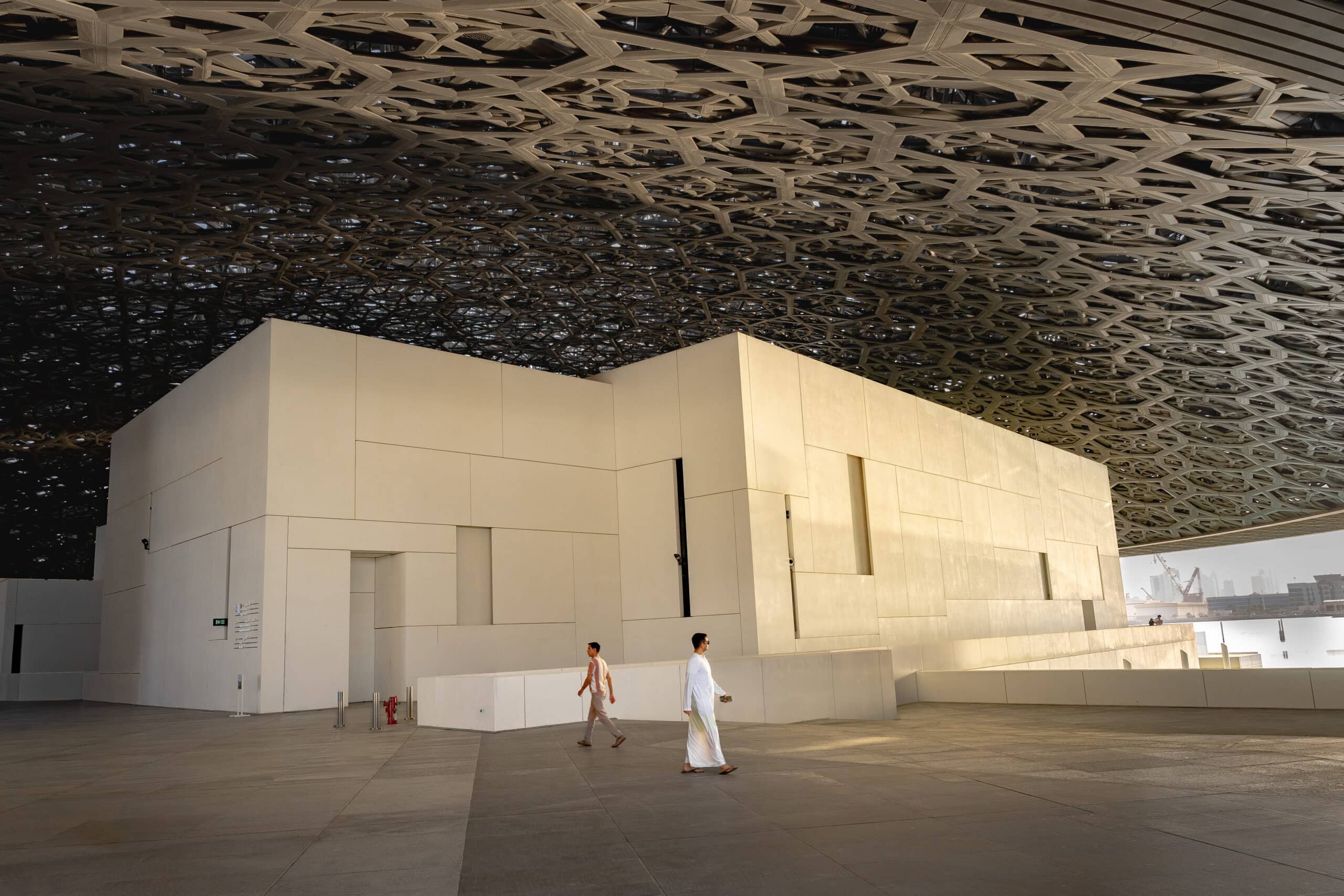 Two men, one wearing traditional Emirati clothes, walk under the Louvre Abu Dhabi dome
