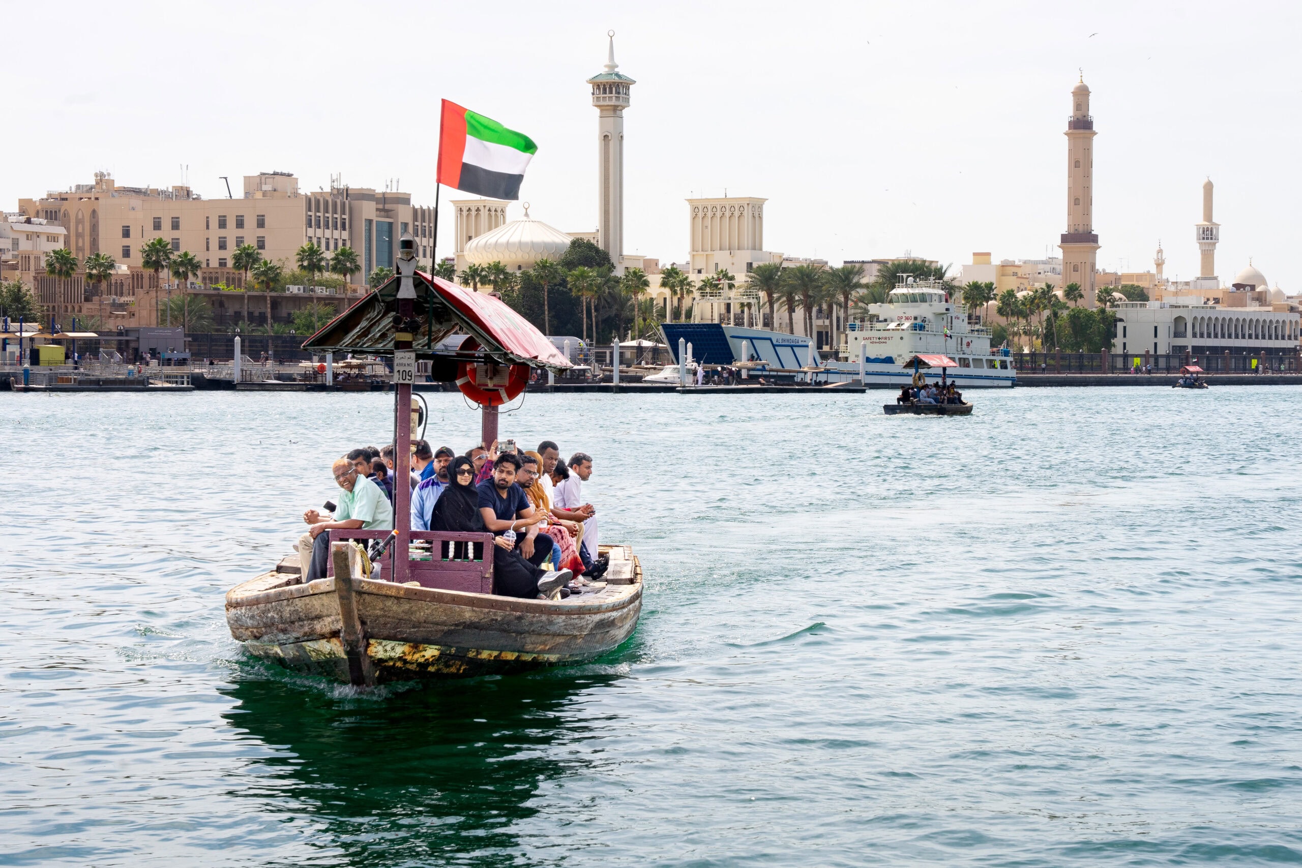 An abra, a small, traditional Emirati boat crosses the Dubai Creek with passengers. You can see tall minarets of Old Dubai in the background