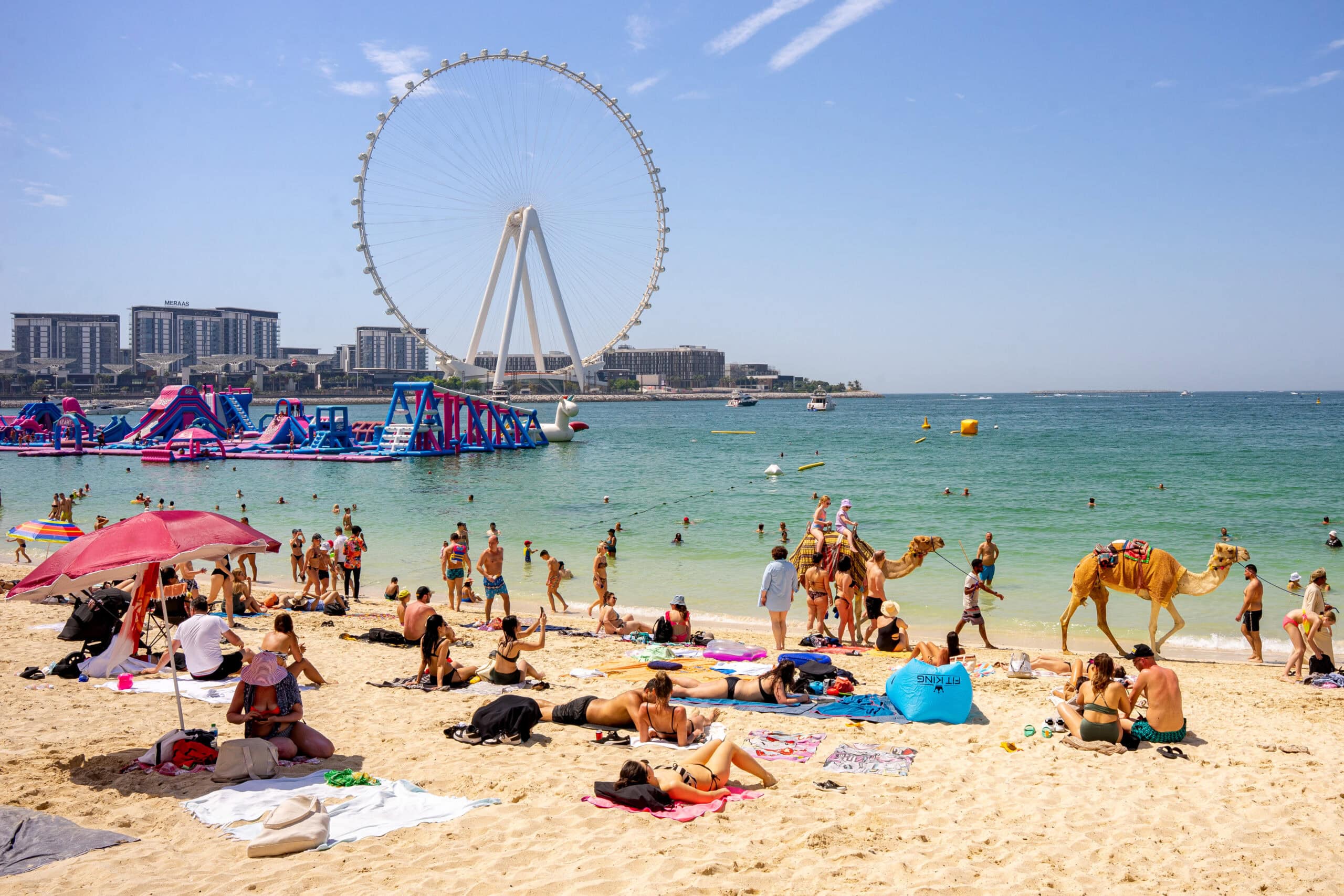 Jumeirah Beach in Dubai. You can see many people sunbathing, there are two camels taking kids for a ride, and the water looks nice and clear