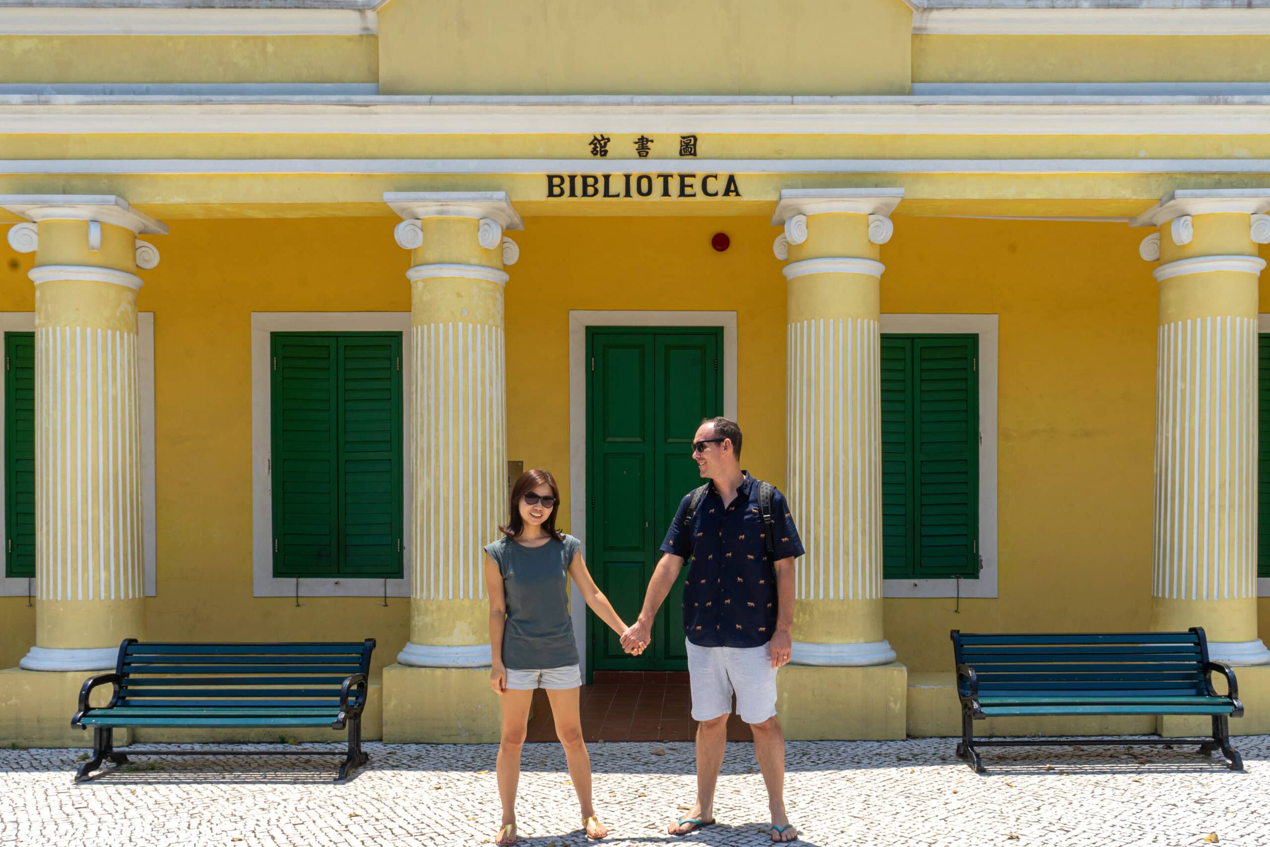 We are posing, hand in hand, in front of a yellow library with green shutters in Coloane Village in Macu. In black letters on the yellow wall you can see the words Bilblioteca and Chinese characters
