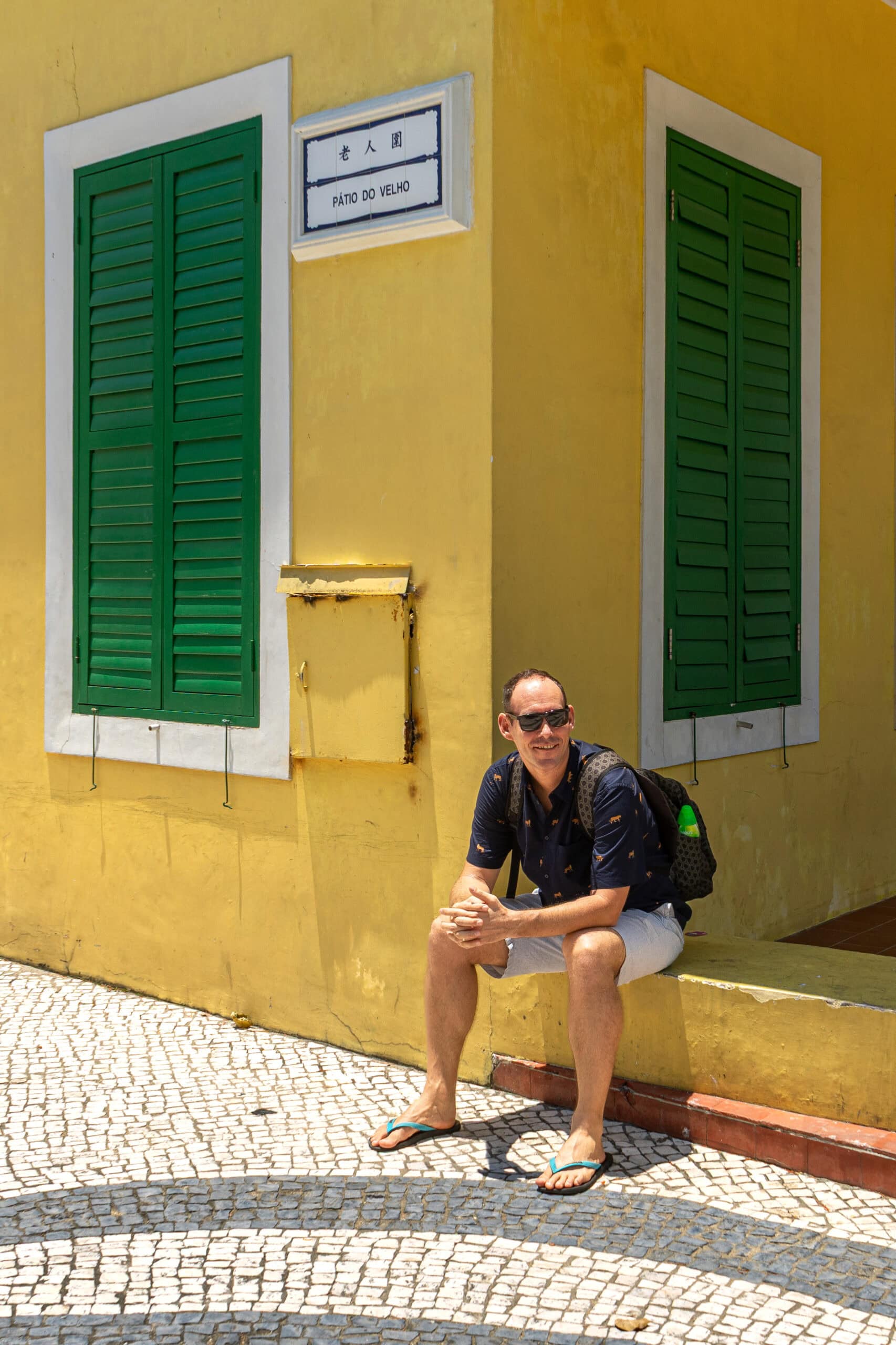I am sitting on a step in front of a yellow colonial era building in Coloane, Macau