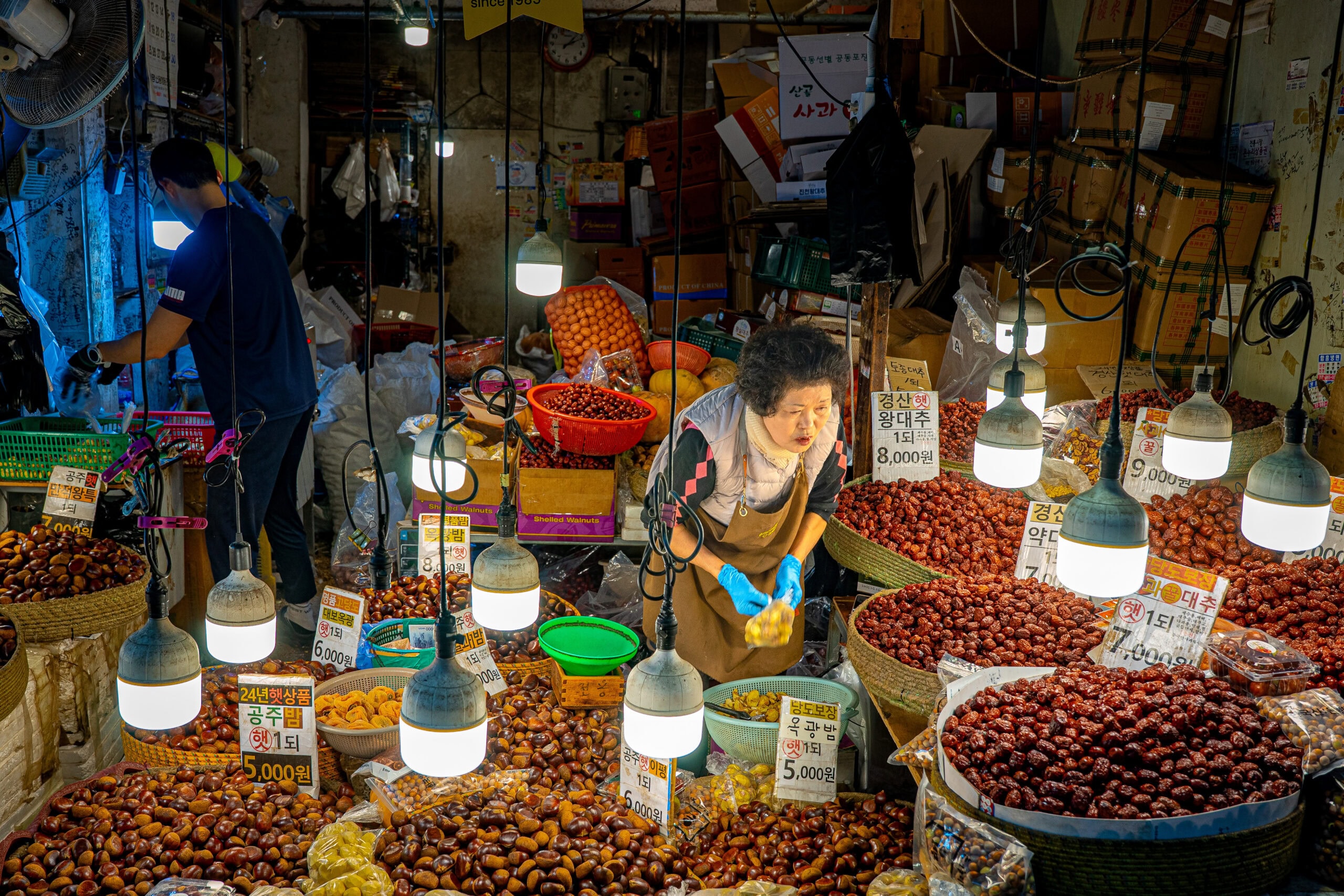 a korean women vendor standing between baskets of jujube and chestnuts at a traditional market in Seoul
