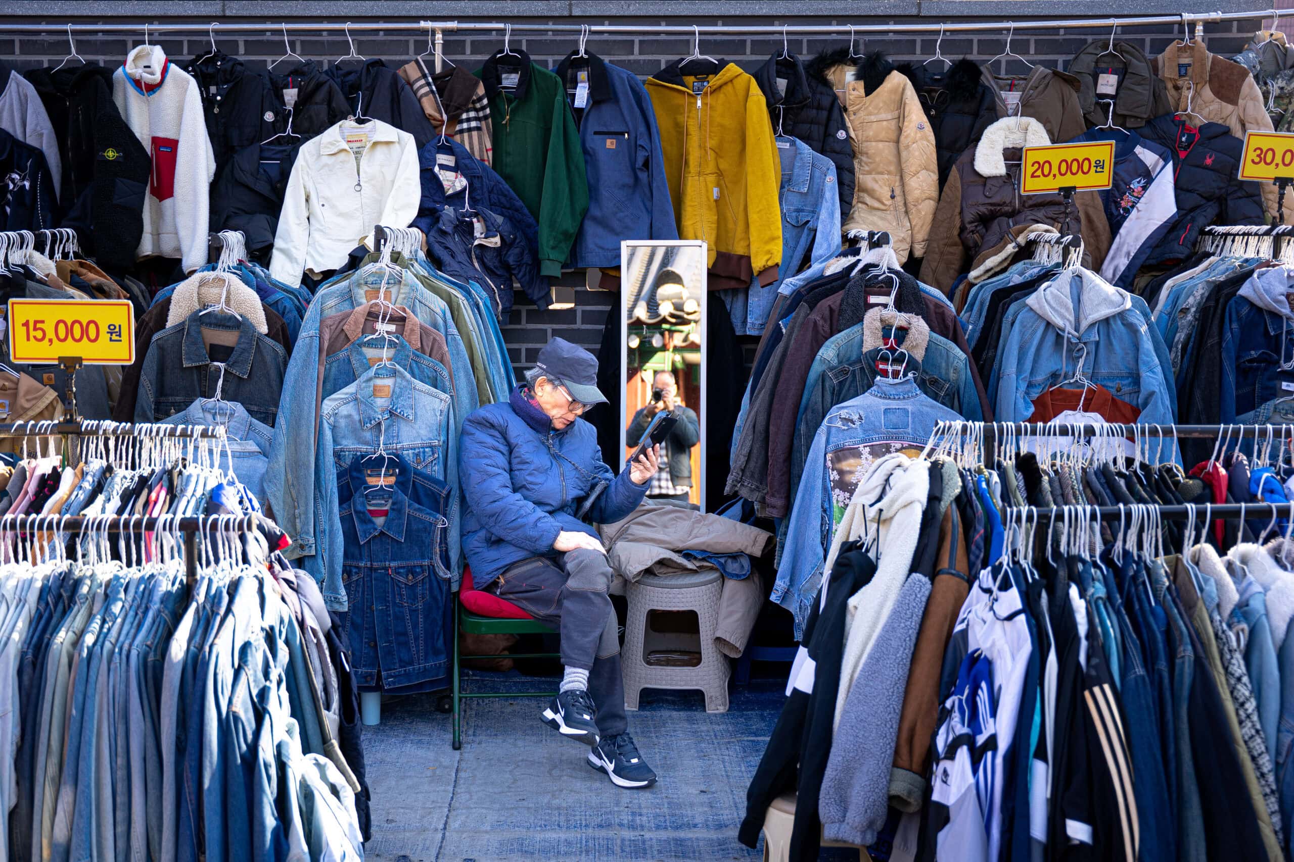 A man sits watching his phone between racks of jean jackets at Dongmyo market