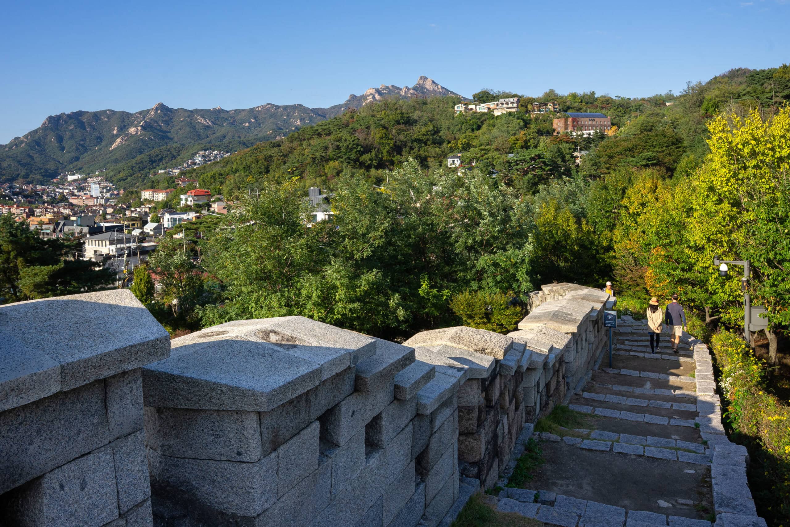 A stone section of the ancient Seoul City Wall snaking down a hillside with hikers walking towards a valley of green trees and distant houses.