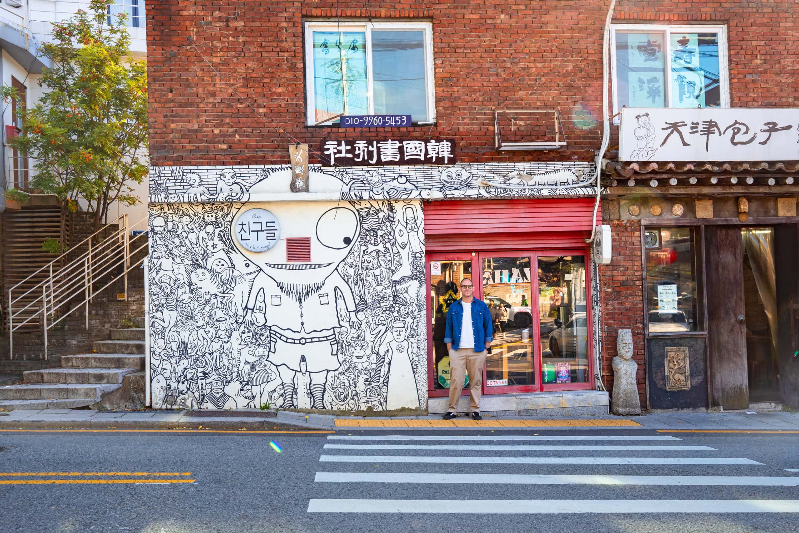 I am standing in front of a red-framed shop window and a wall covered in intricate black and white street art in Buam-dong