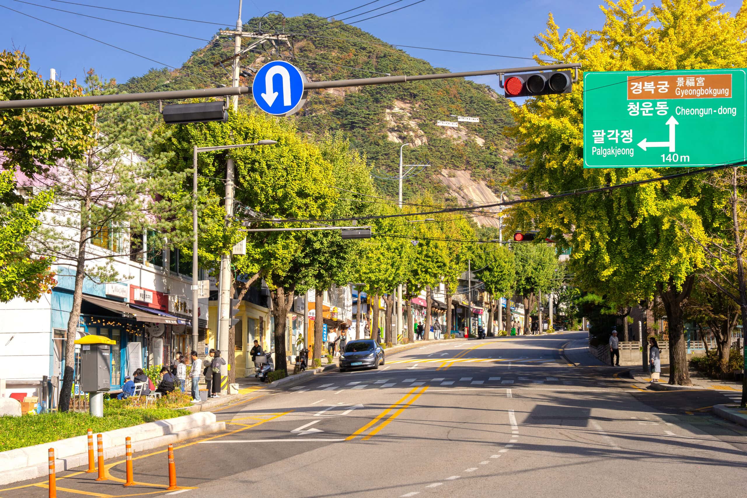 The main street in Buam-dong, Seoul lined with vibrant yellow ginkgo trees in autumn with mountain peaks visible in the distance under a blue sky.