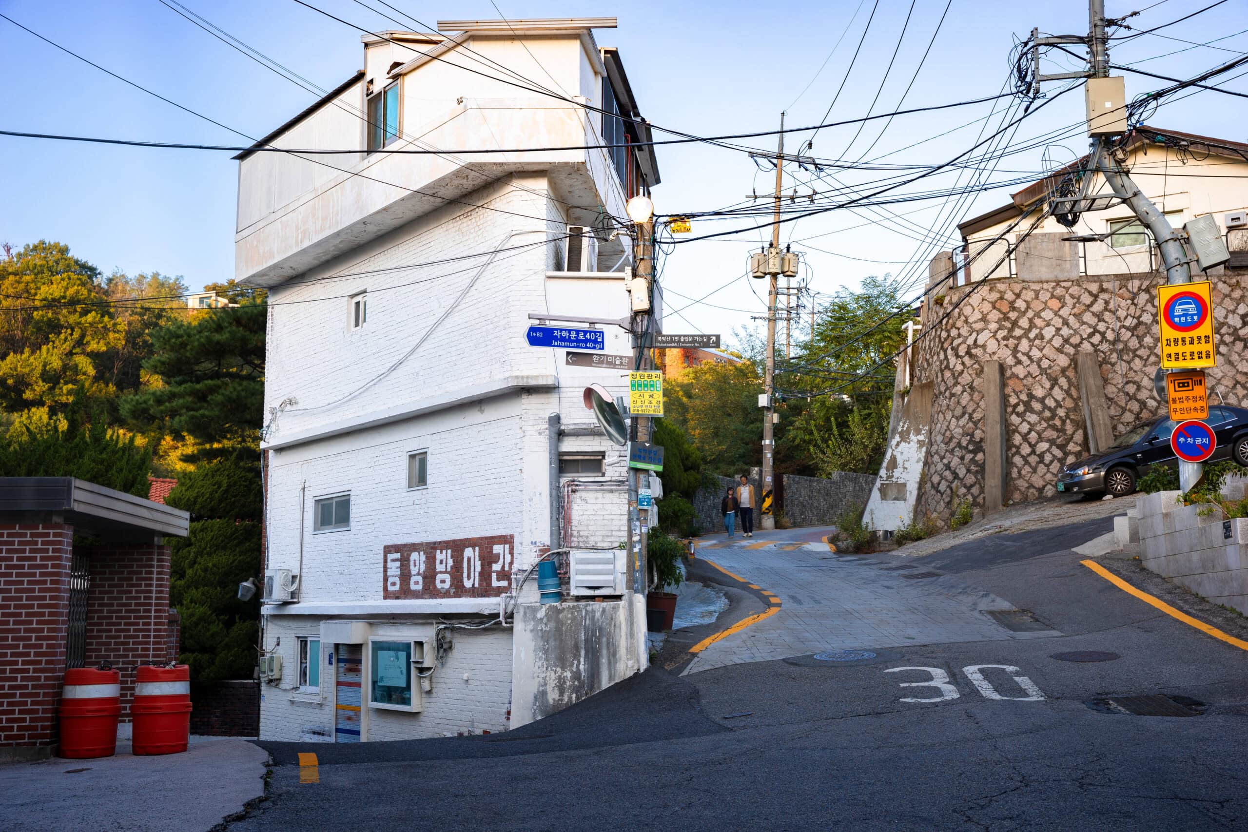 A narrow, winding asphalt road in Seoul leading past a unique white multi-storey building with various street signs and a stone retaining wall.