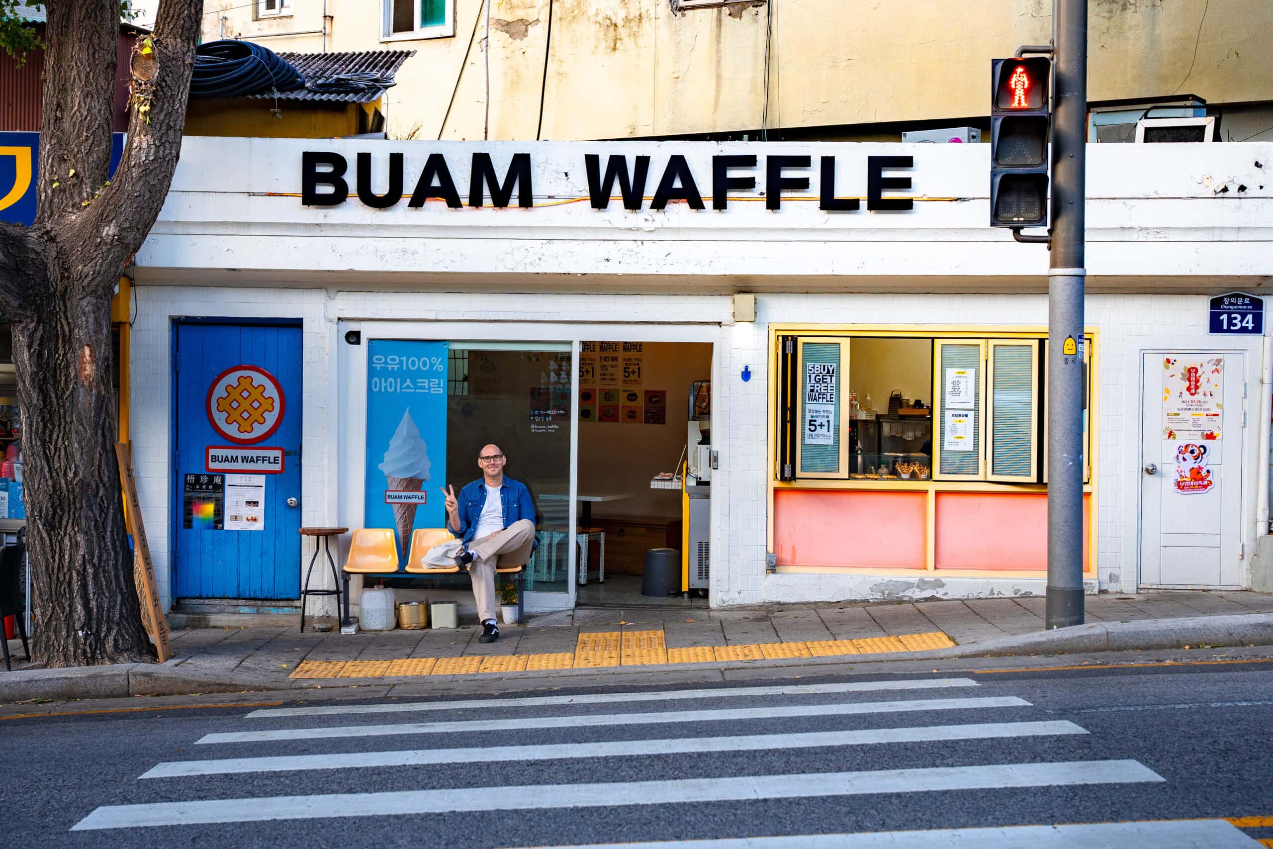 I am sitting on a yellow chair outside the Buam Waffle shop in Seoul with a blue door and a vintage aesthetic.
