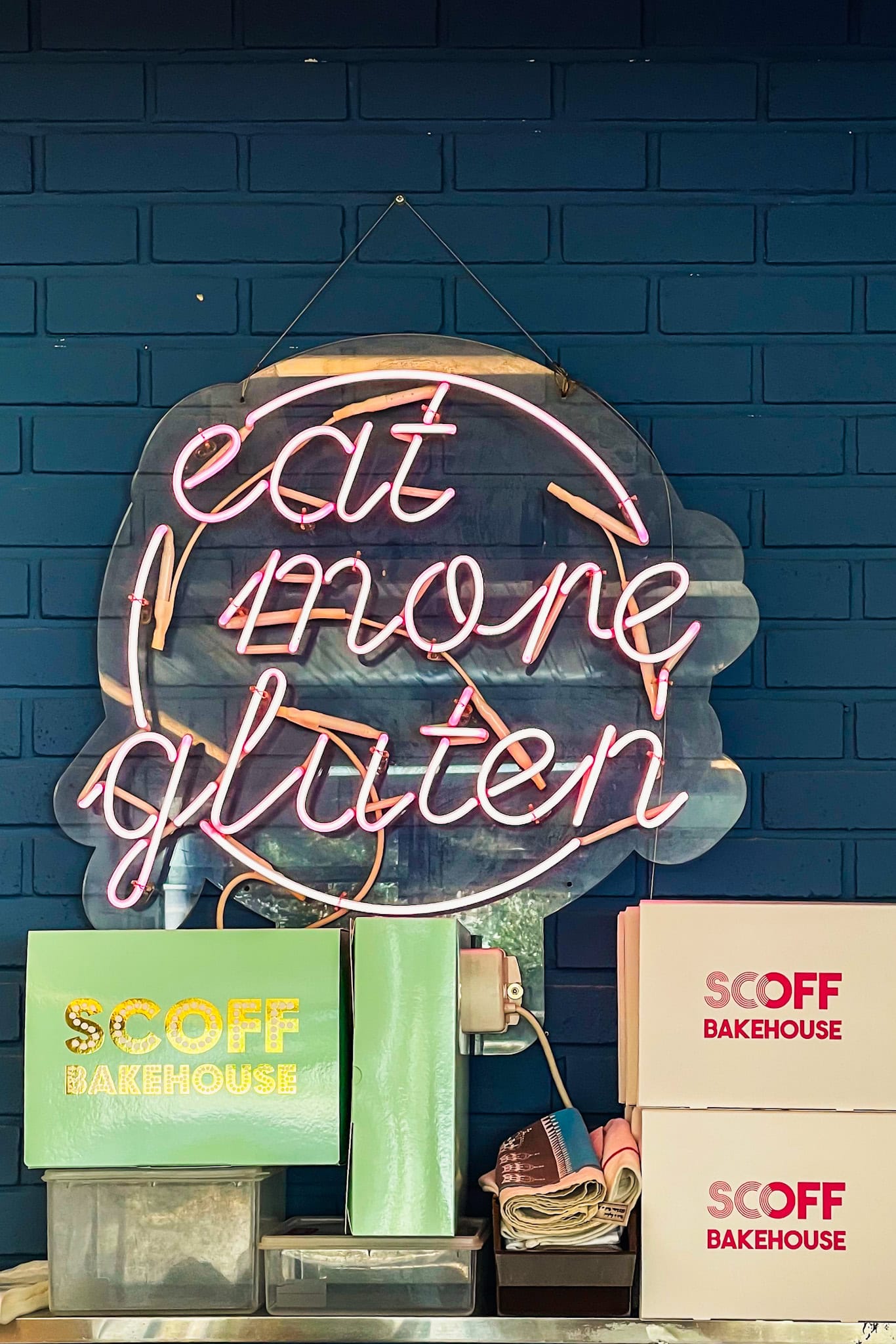 A pink neon sign hanging above take-out boxes from Scoff Bakehouse. It reads: Eat more gluten