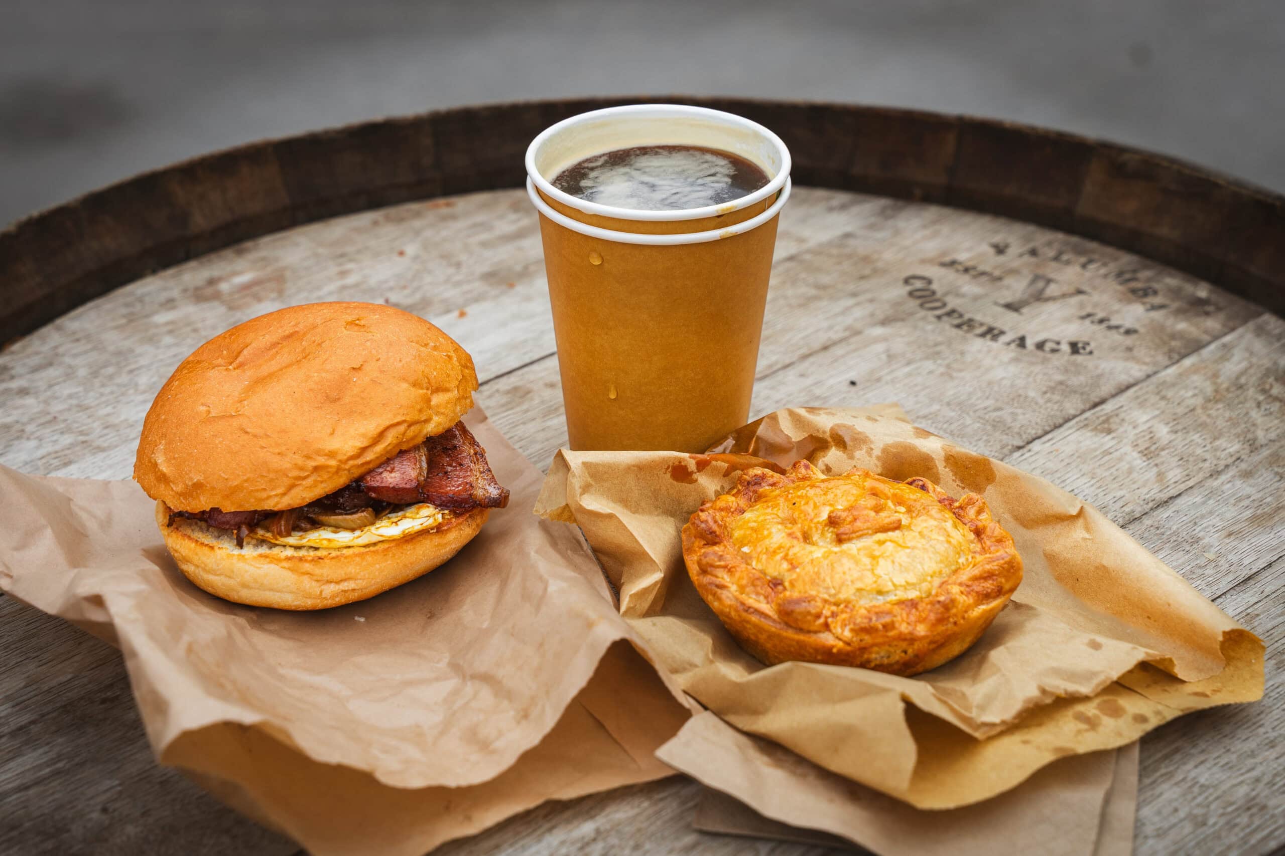 Our breakfast we had at Barossa Farmer's market: a bacon and egg breakfast bun, a beef and cheese pie, and a paper cup of coffee, all sitting on a wooden wine barrel
