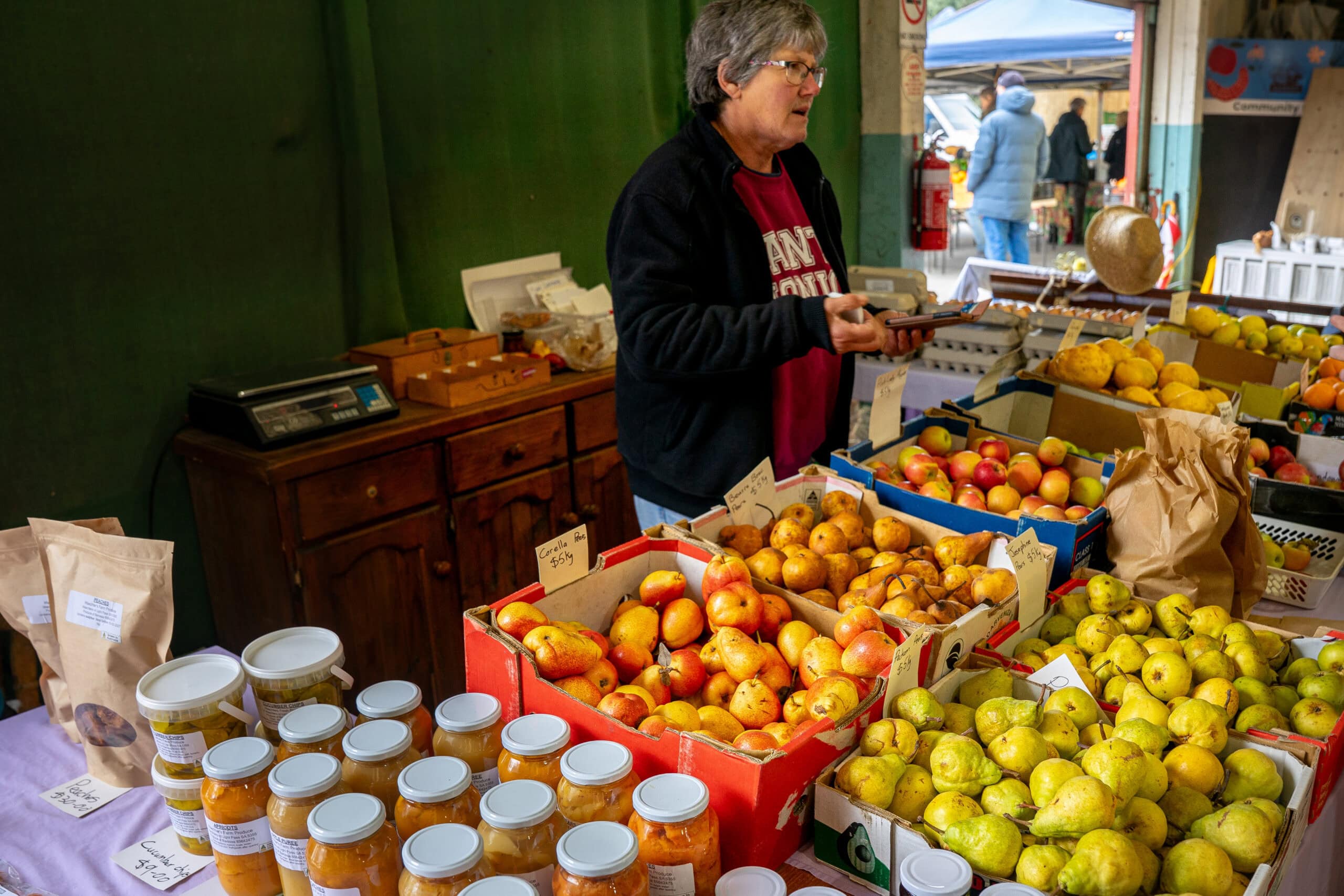A stall holder stands behind displays of apples, pears, and jars of preserves at the Barossa Farmers Market
