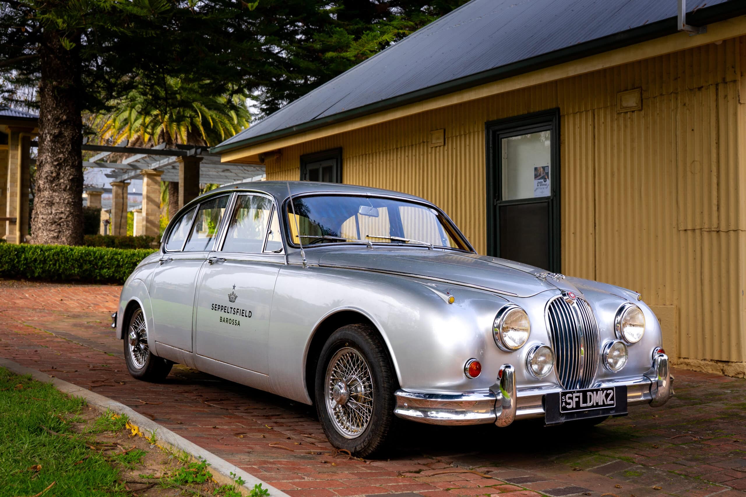 A vintage, silver Jaguar Mark 2  with Seppeltsfield Barossa branding on the door, parked in front of a yellow corrugated iron building