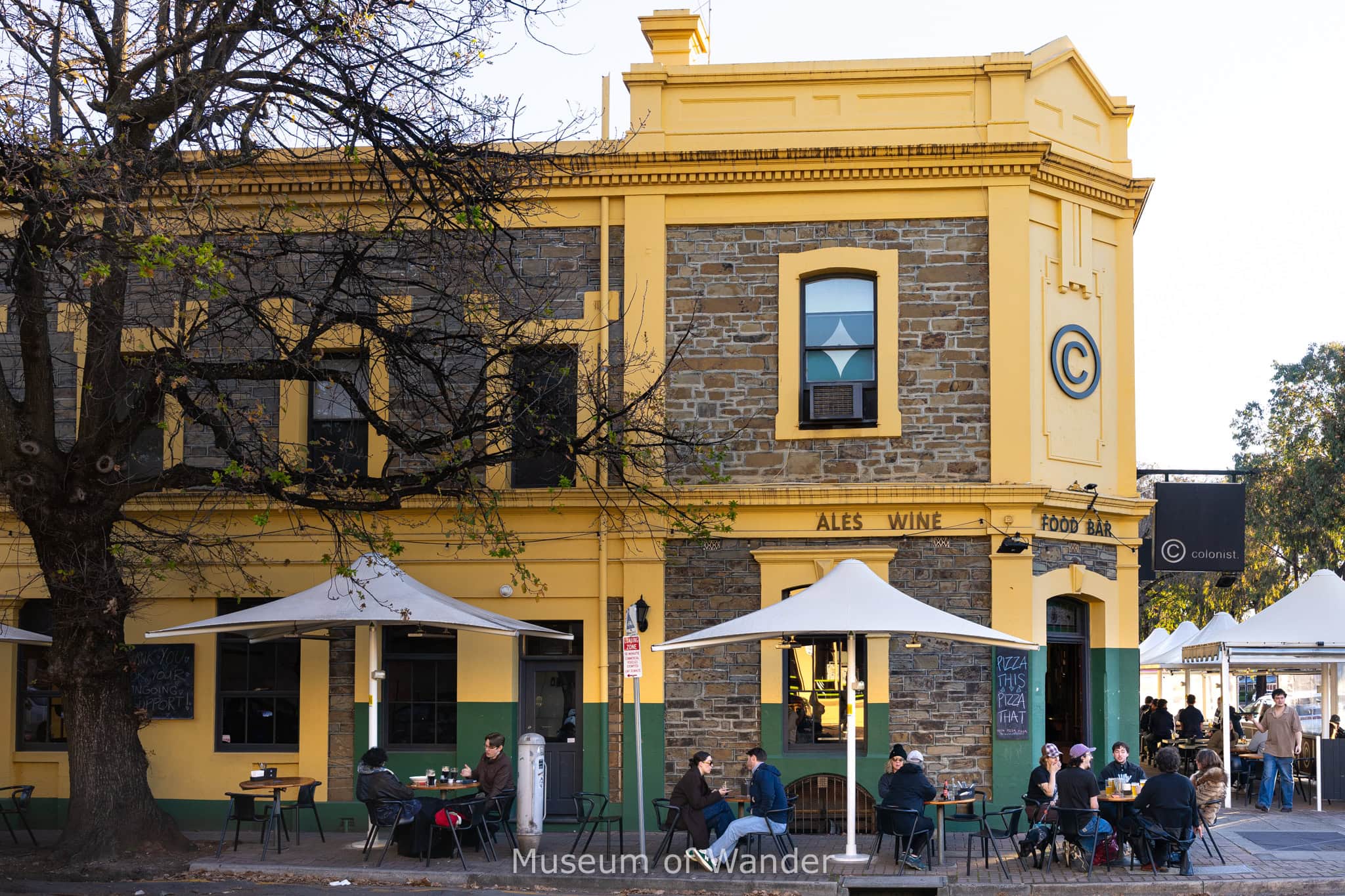 People sitting outside a yellow and green pub with stone walls drinking beer on a sunny winter afternoon in Adelaide, South Australia