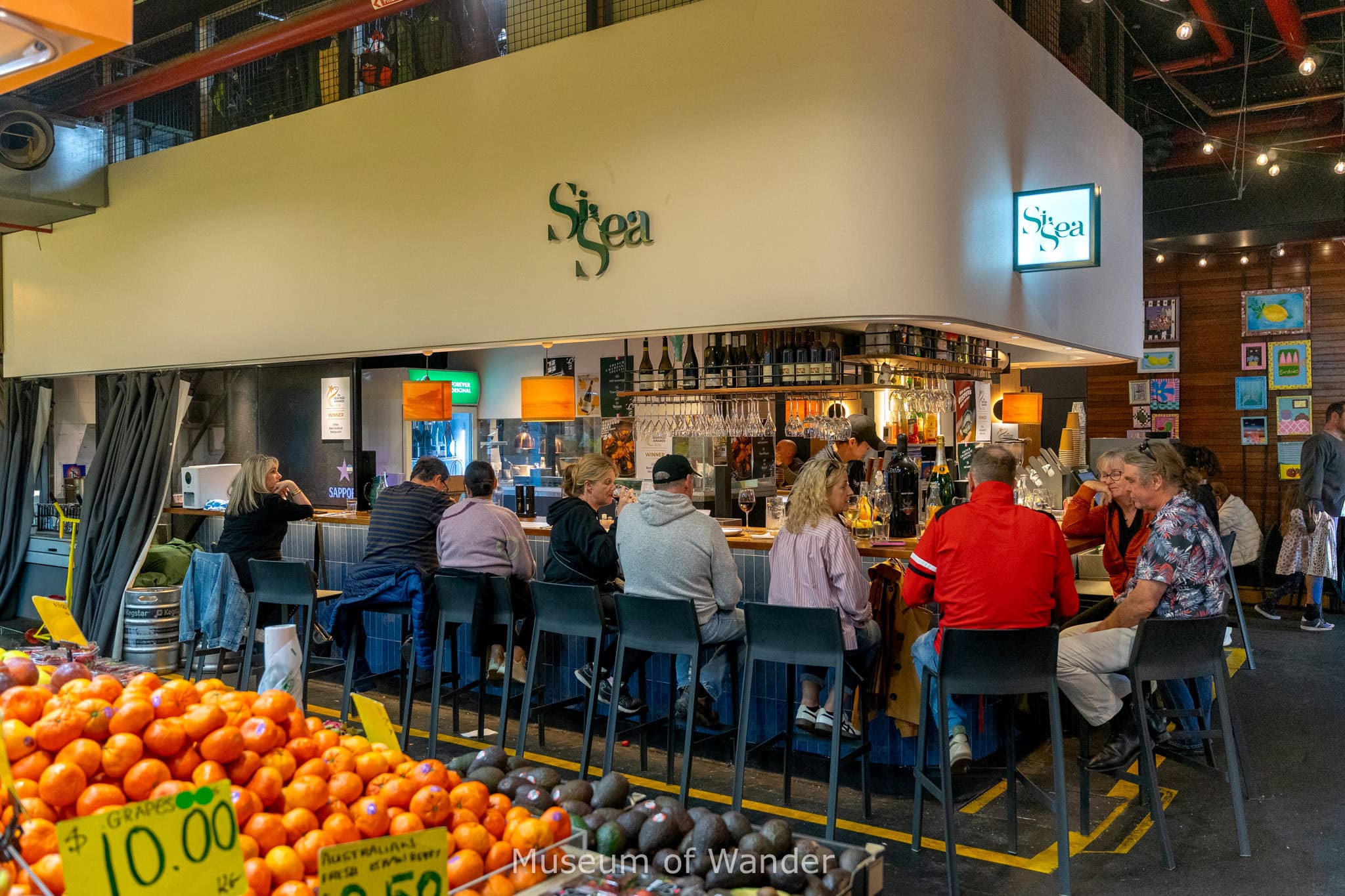 A row of diners sit at the counter of Si Sea, the best seafood restaurant in South Australia, inside Adeliade Central Market. You can see displays of oranges and avocados from a fruit stall in the foreground