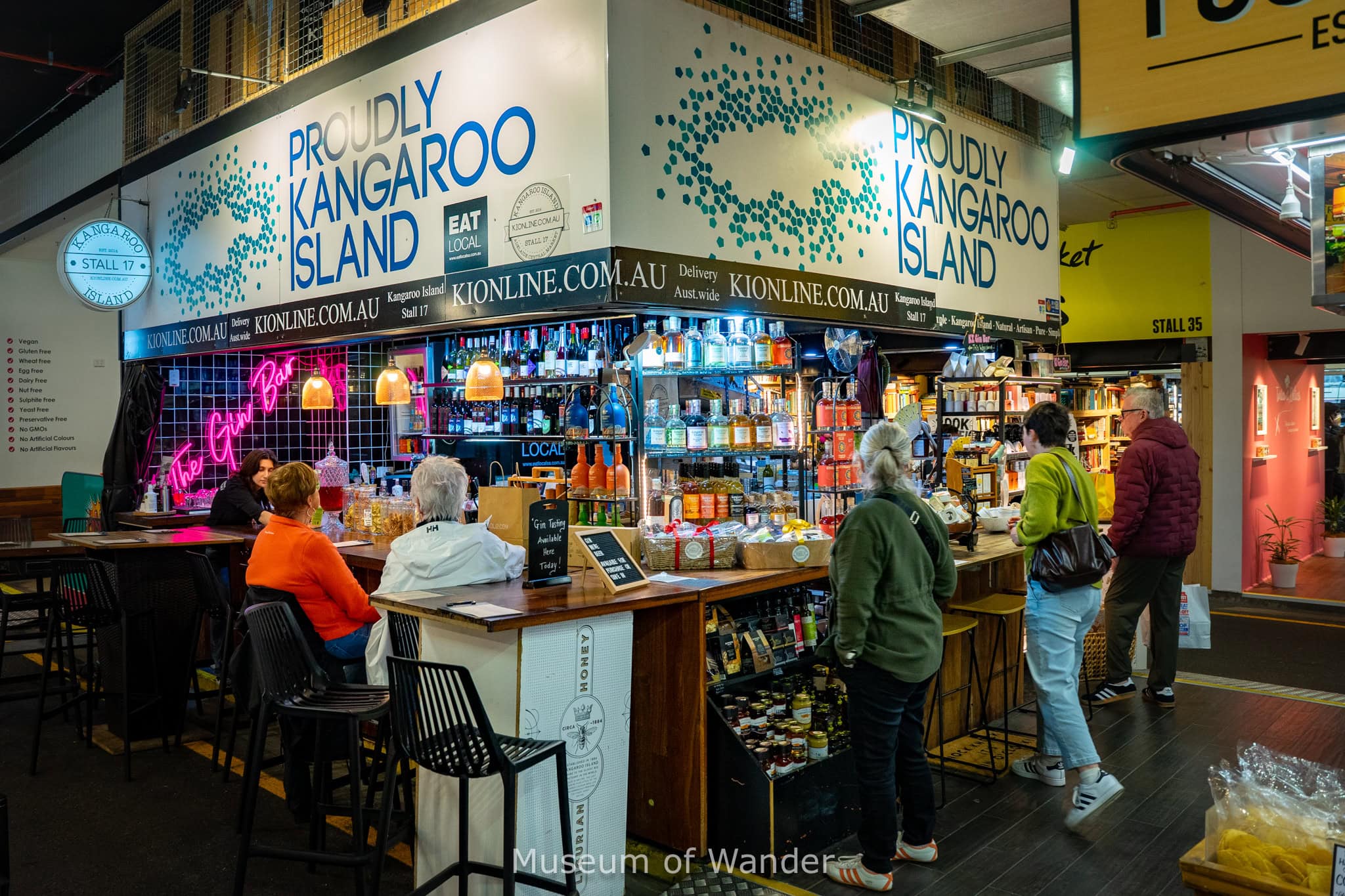 People sitting or standing around the small bar of the Kangaroo Island stall. There are bottles of wine and gin as well as other organic produce for sale