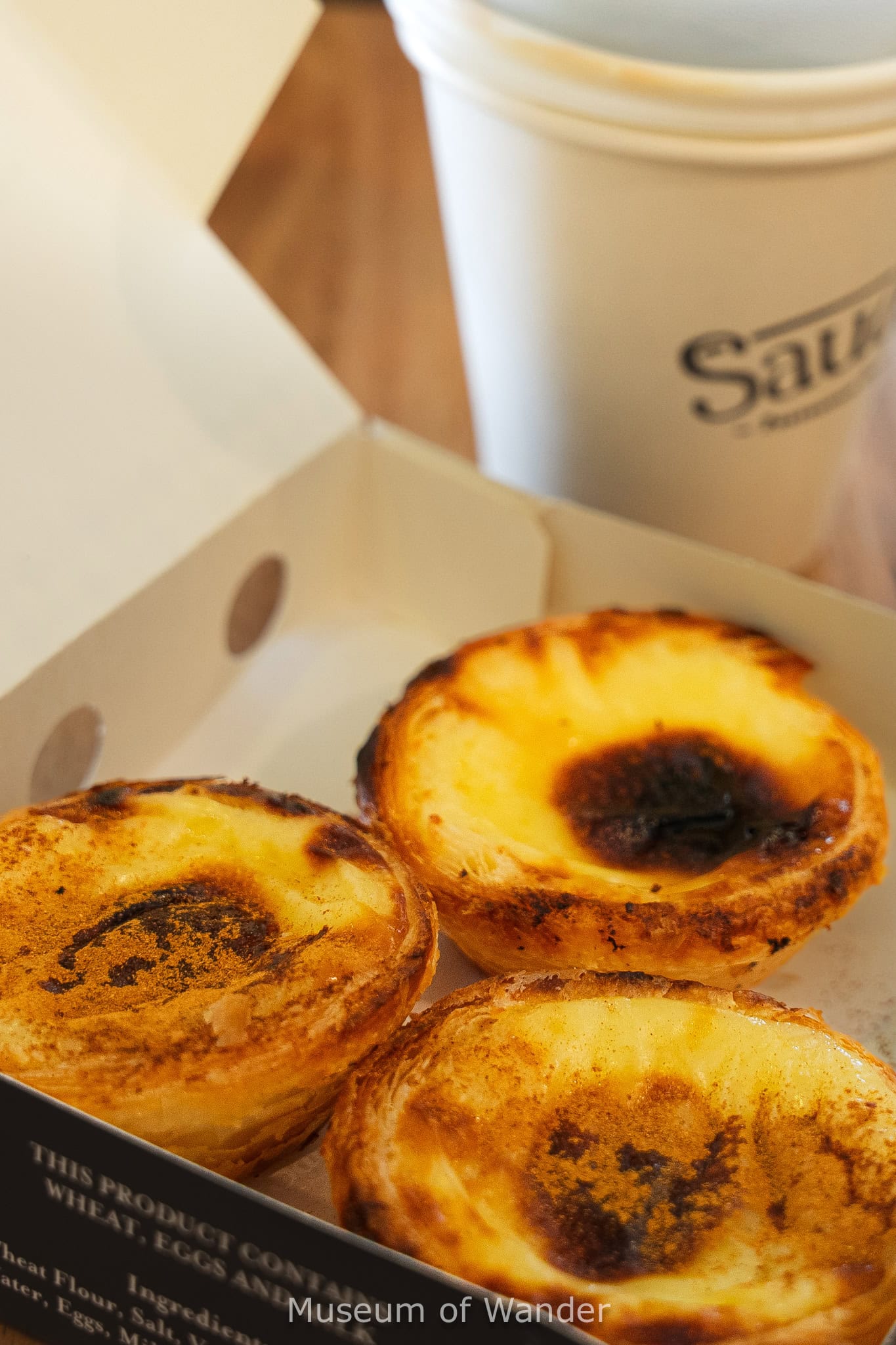 Three golden egg tarts dusted with cinnamon in a white paper box with a take away coffee from Saudade at the Central Market in Adelaide